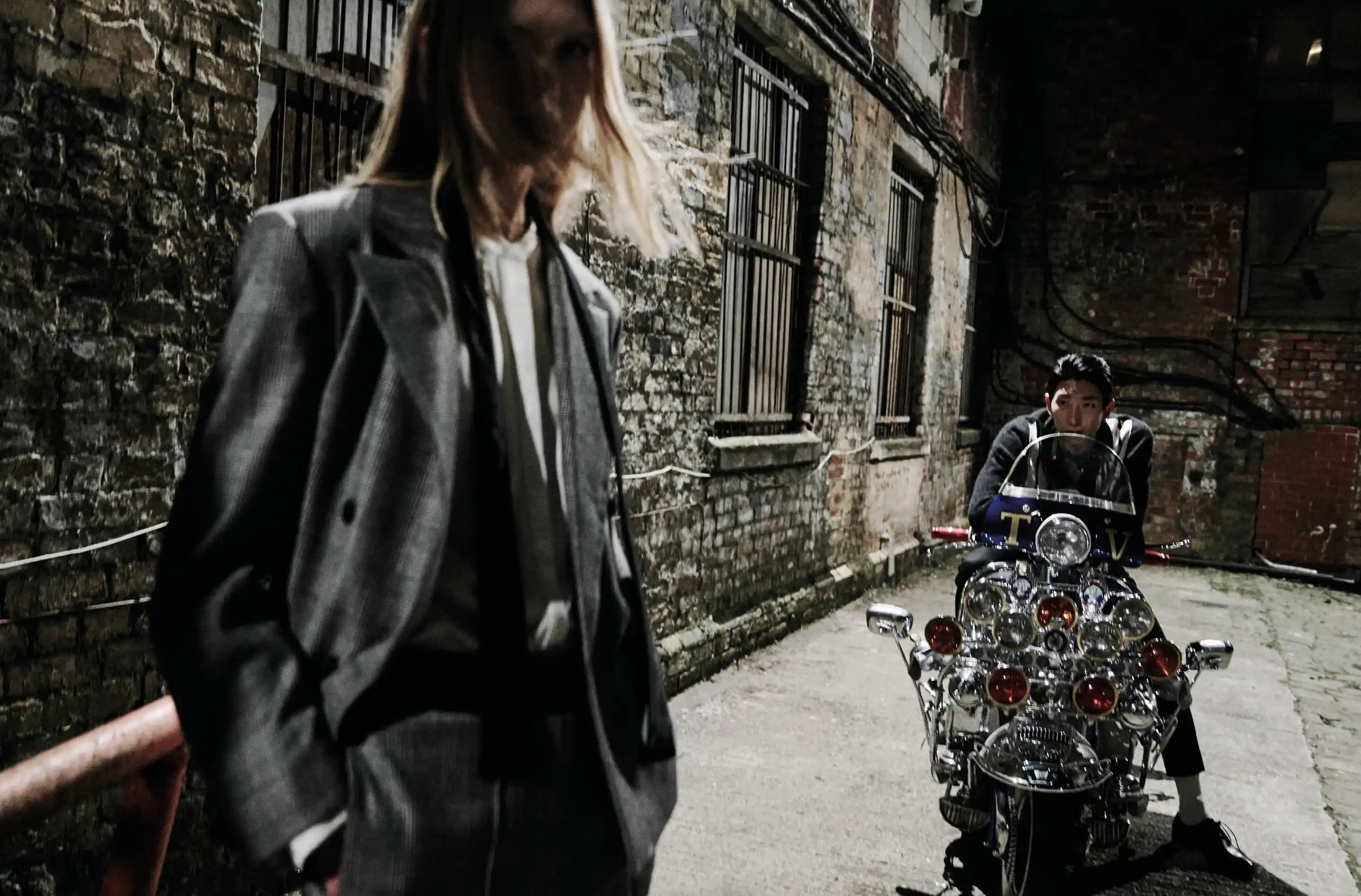 A woman wearing a tailored coat standing in the foreground, a man sitting on a motorcycle in the background.