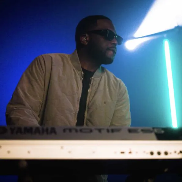 A band-member playing the keyboard wearing a beige bomber and black sunglasses.