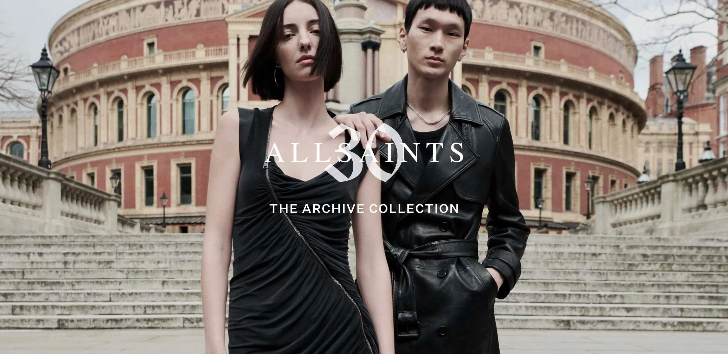 A male and female model standing in from of the Royal Albert Hall wearing a black asymmetrical dress and leather trench coat