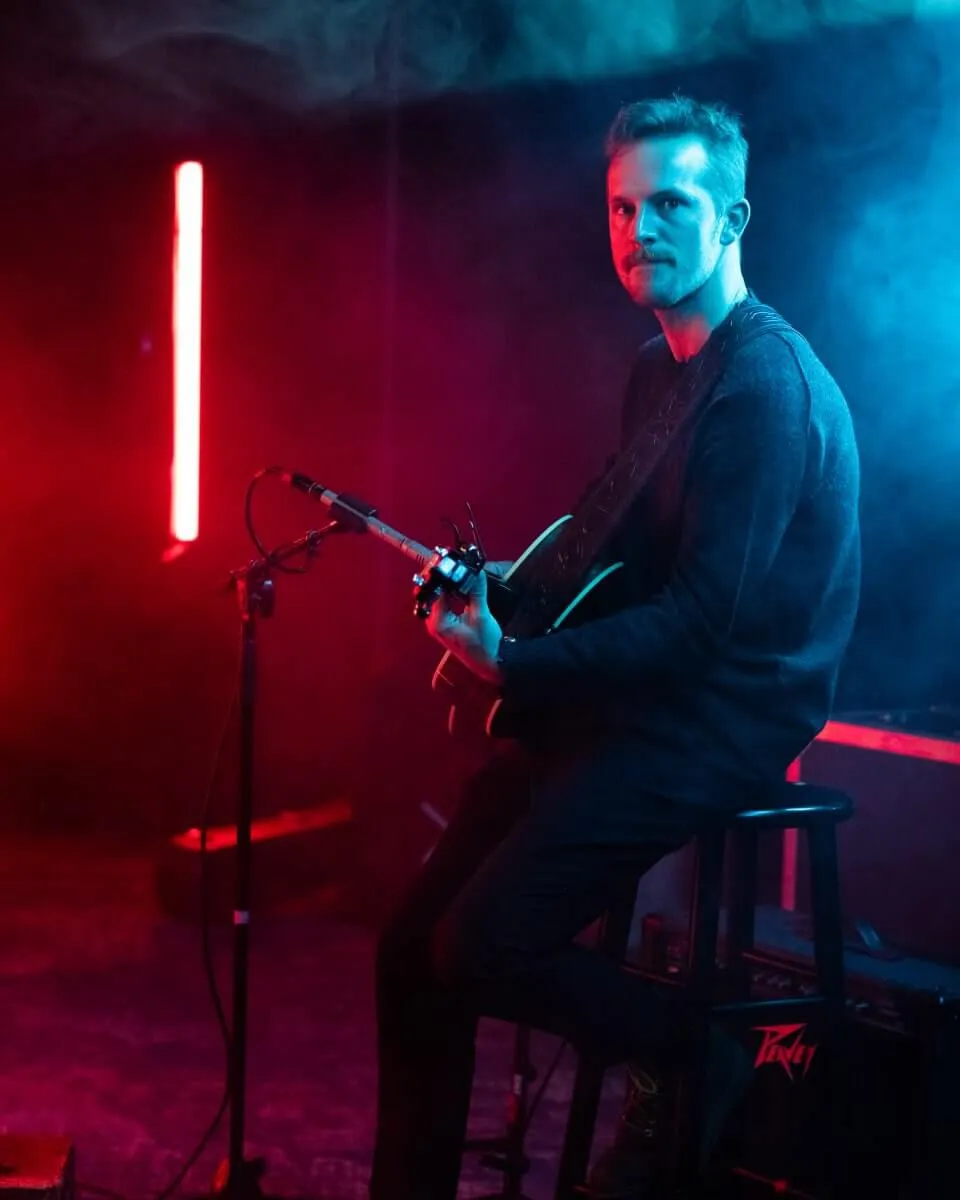 A guitarist playing in front of a microphone, with red and turquoise light.