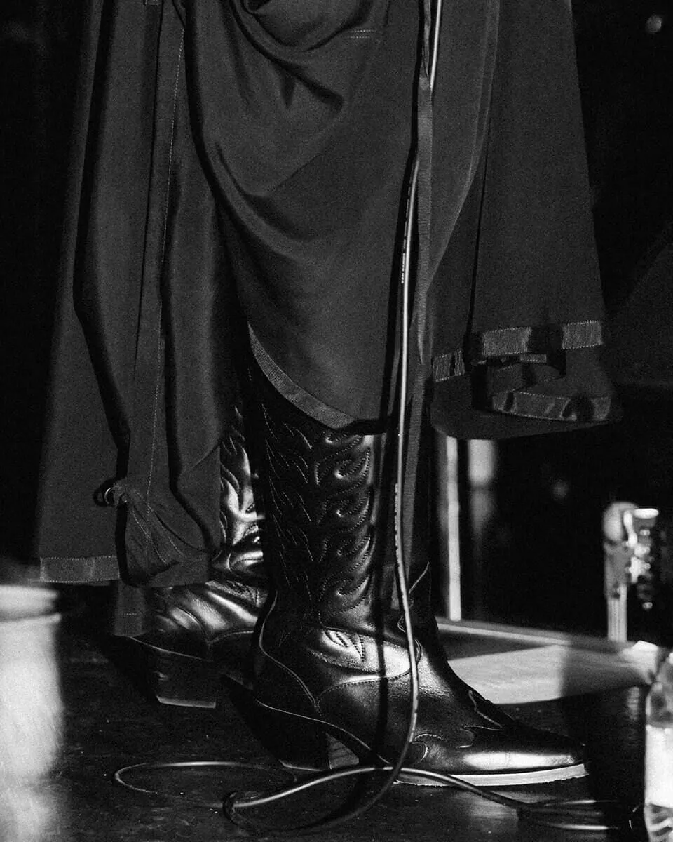 Black and white shot of Etta Marcus standing on stage, her cowboy boots and bottom of her dress are visable.