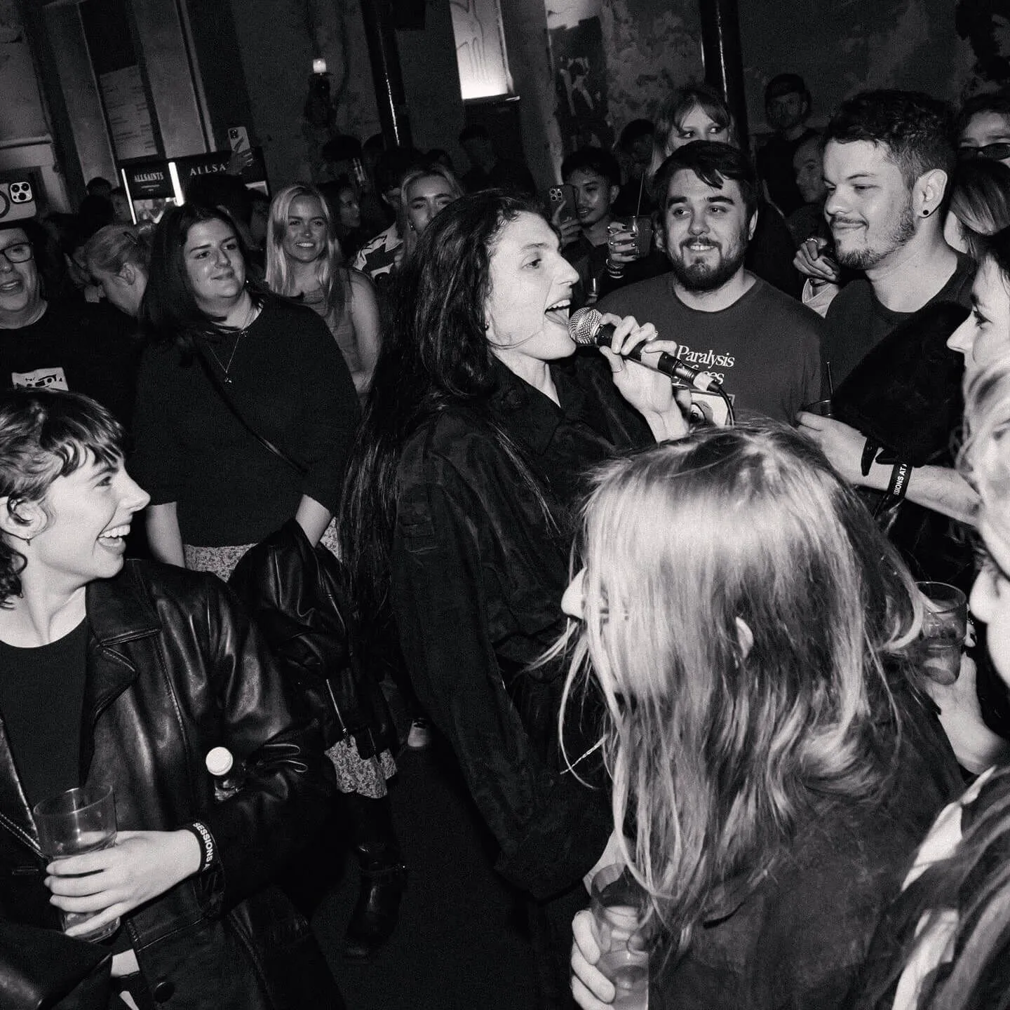 Black and white shot of Lucia & The Best Boys front singer singing into a microphone standing in a crowd of people..