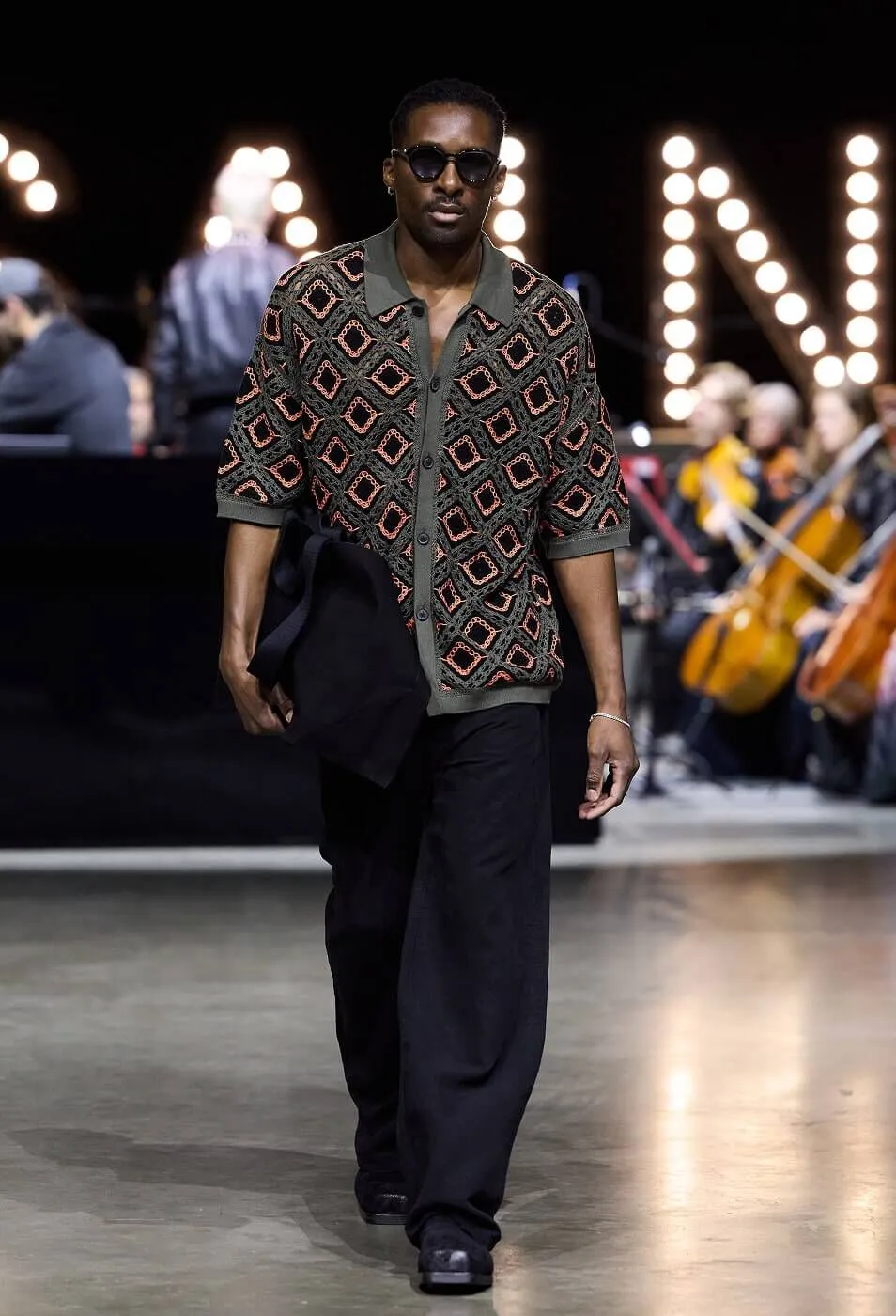 Man wearing patterned button up shirt with black trousers and a black bag