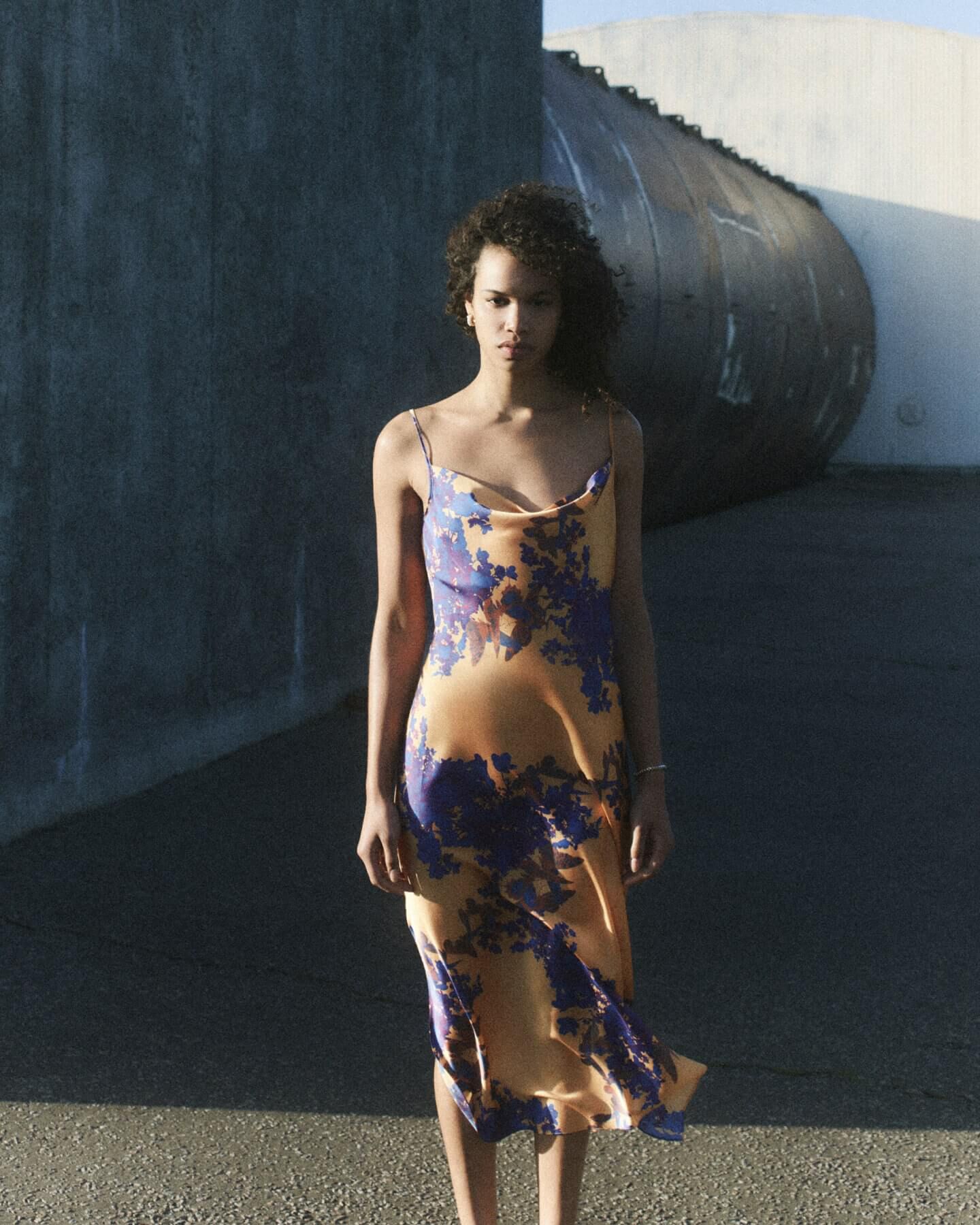 Woman wearing an orange slip dress with purple florals standing in front of a concrete structure