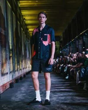 A man wearing a short sleeved black shirt with a bold print on it with black shorts catwalking.