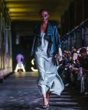 A woman wearing a silver shiny dress with a leather jacket catwalking.