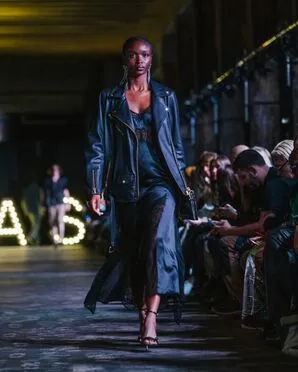 A woman wearing an black silk slipdress with lace inlays and a black leather jacket catwalking.