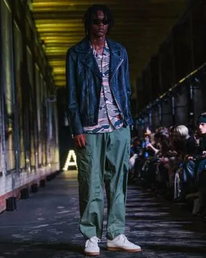 A man wearing a black leather jacket over a camo shirt and khaki military style trousers catwalking.