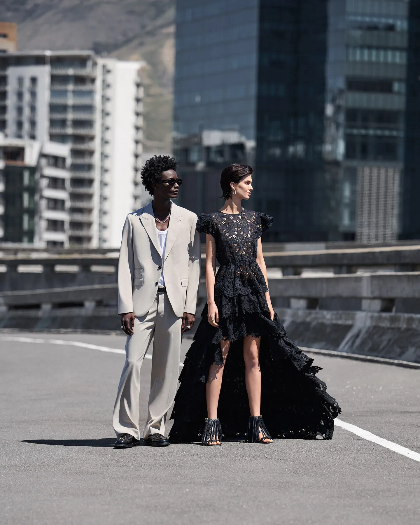 Photograph of male model in beige next to a female model in a blakc dress on a road.