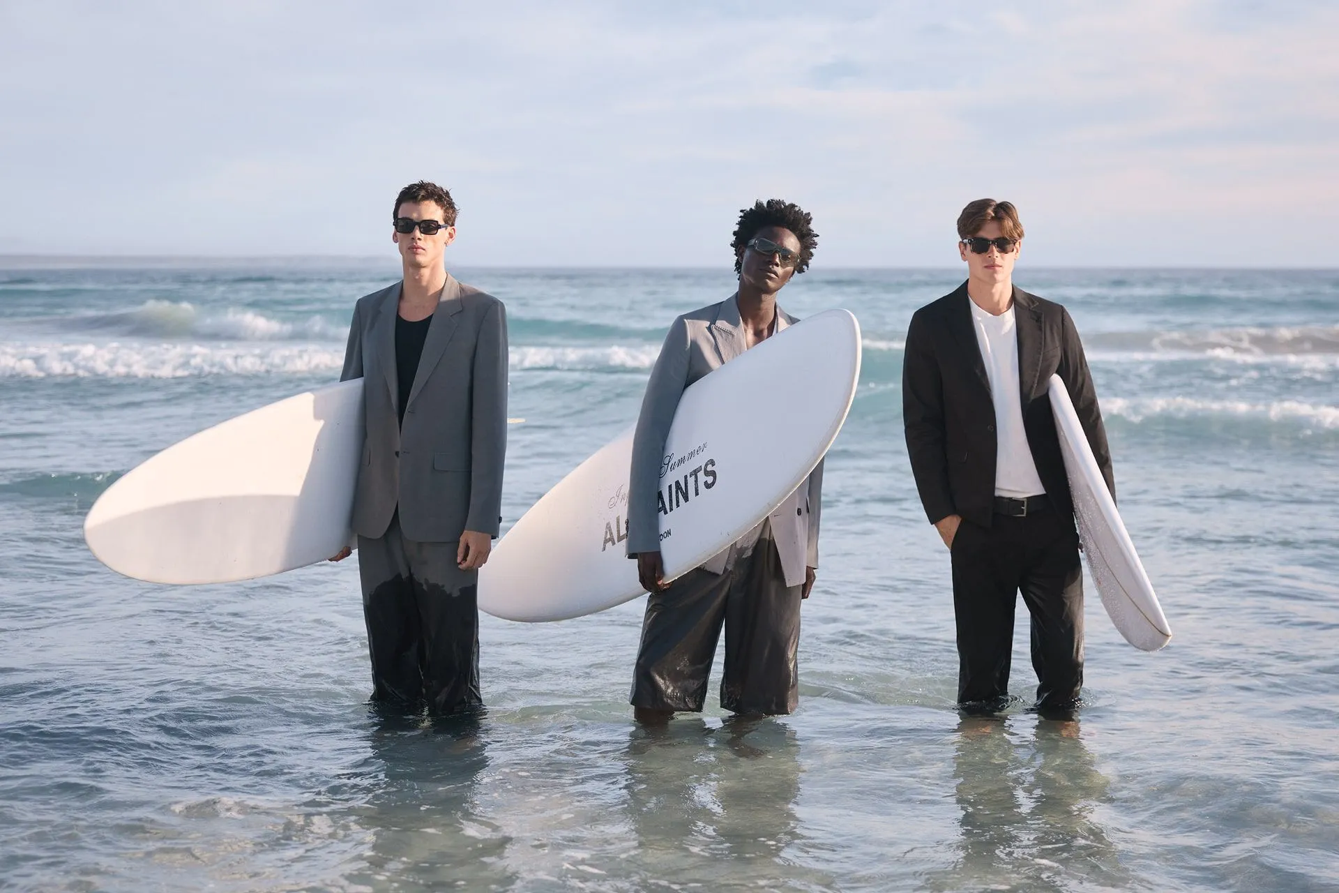 Photograph of three male models in suits on the beach holding surfboards.