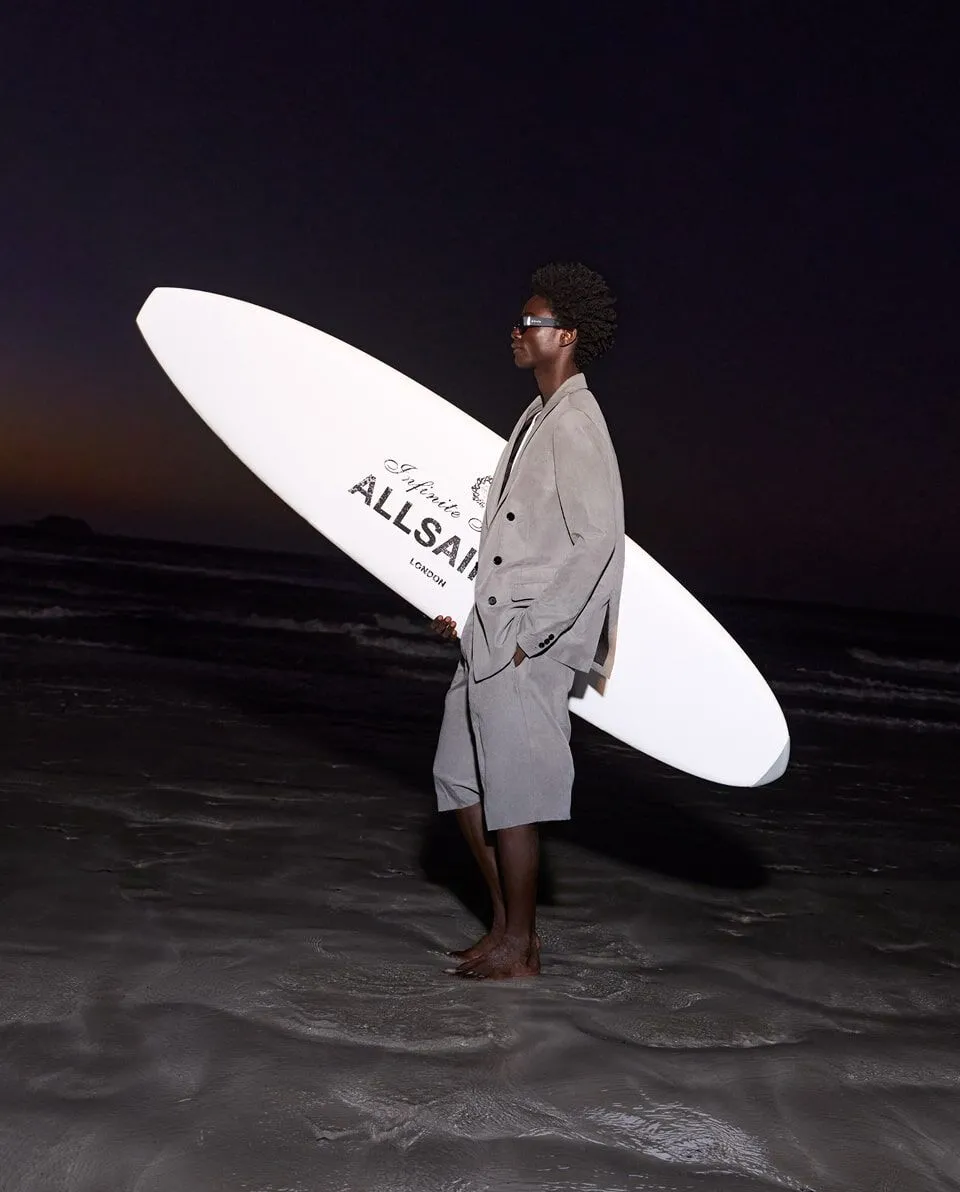 Photograph of a male model in a suit and holding a surfboard on the beach.