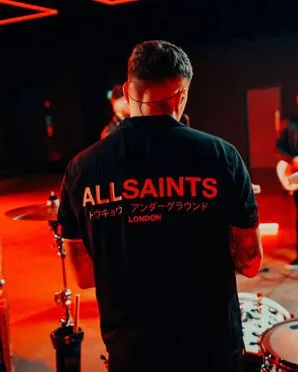 Performer stands with back turned and wears AllSaints branded short sleeved Shirt..