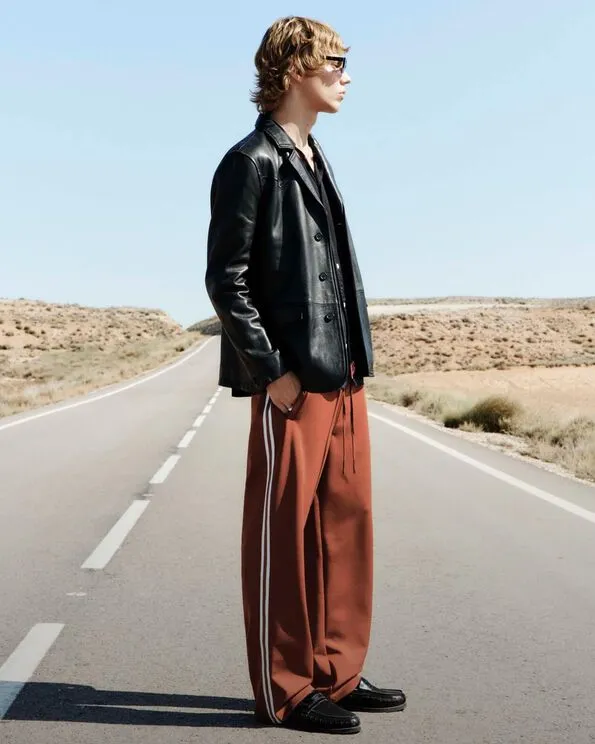 Man wearing sienna coloured sweatpants, a black leather jacket and black leather shoes standing on a road.