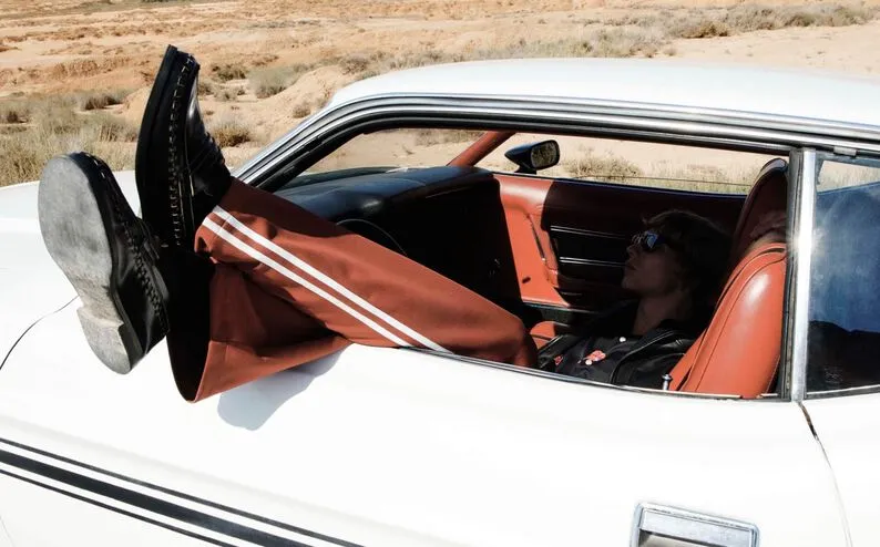 Photograph showing a model resting in an old fashioned white car wearing sienne sweatpants and black leather loafers hanging out of the window.