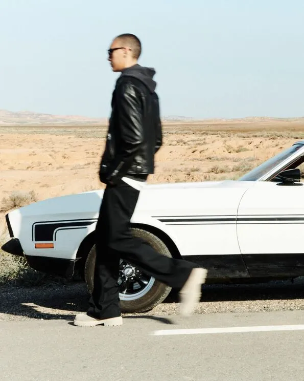 Portrait of a model wearing a black leather jacket with a hoodie, black trousers and sunglasses walking past a white vintage car.