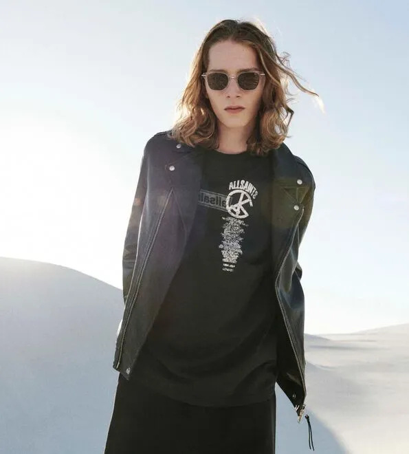 A man wearing sunglasses and an open black leather jacket over a black graphic t-shirt, standing in the desert.