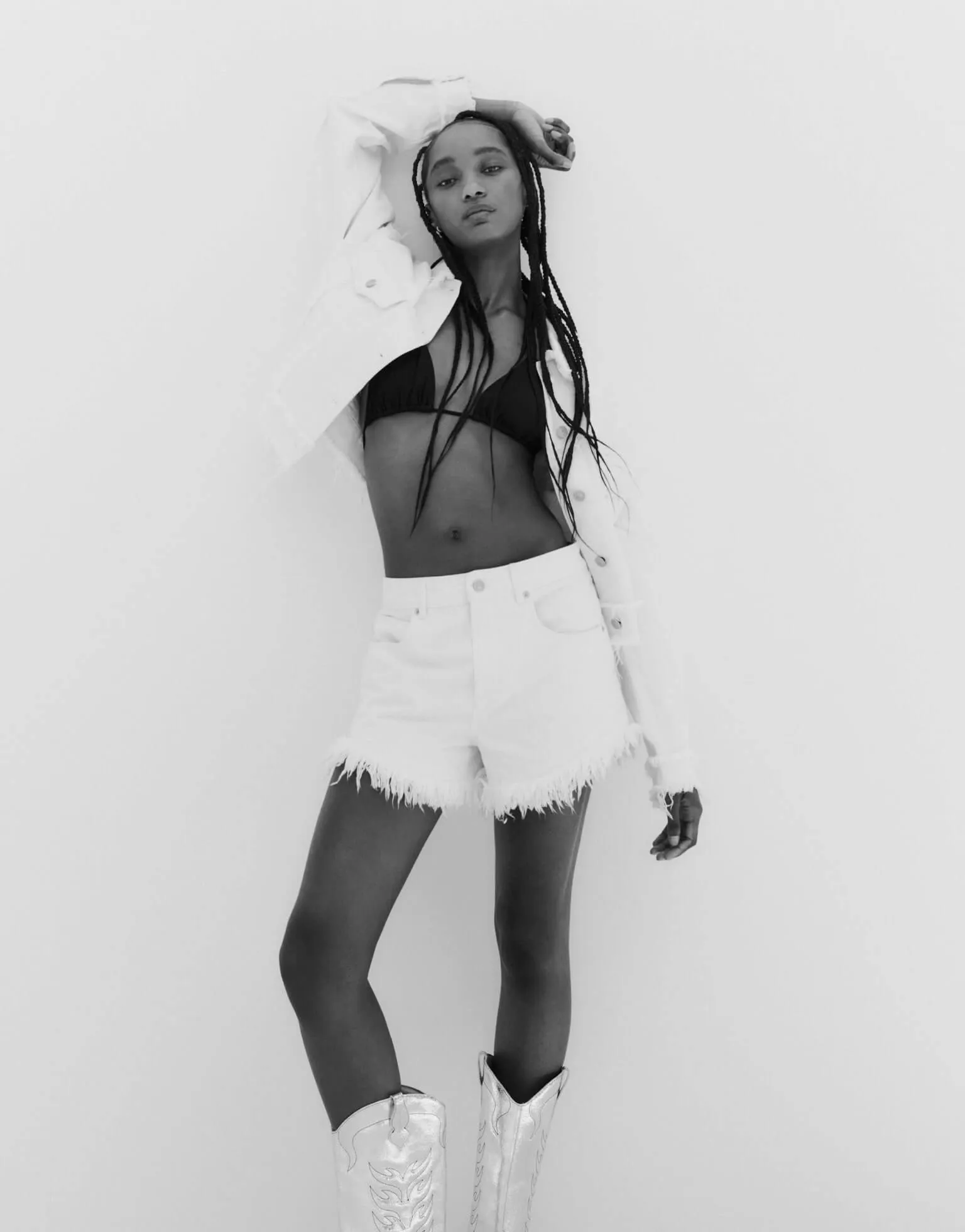 Photograph showing a female model wearing a black bikini underneath a white denim jacket, and white raw edged denim shorts.