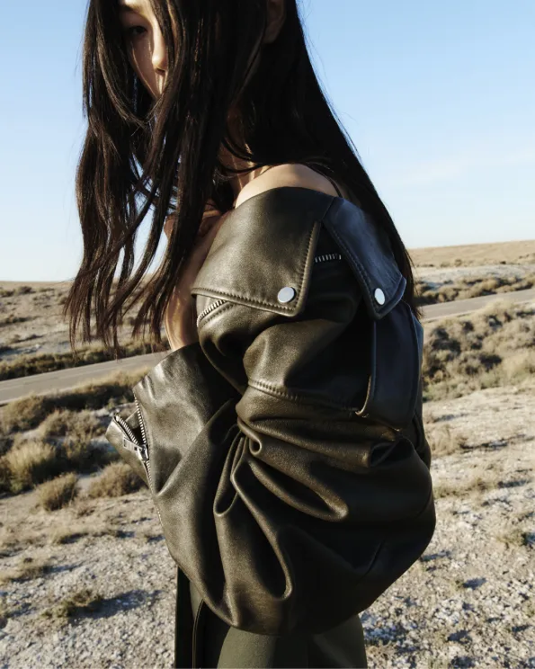 A profile photograph of a model wearing a fringed black leather jacket with black wide leg jeans standing in the desert.