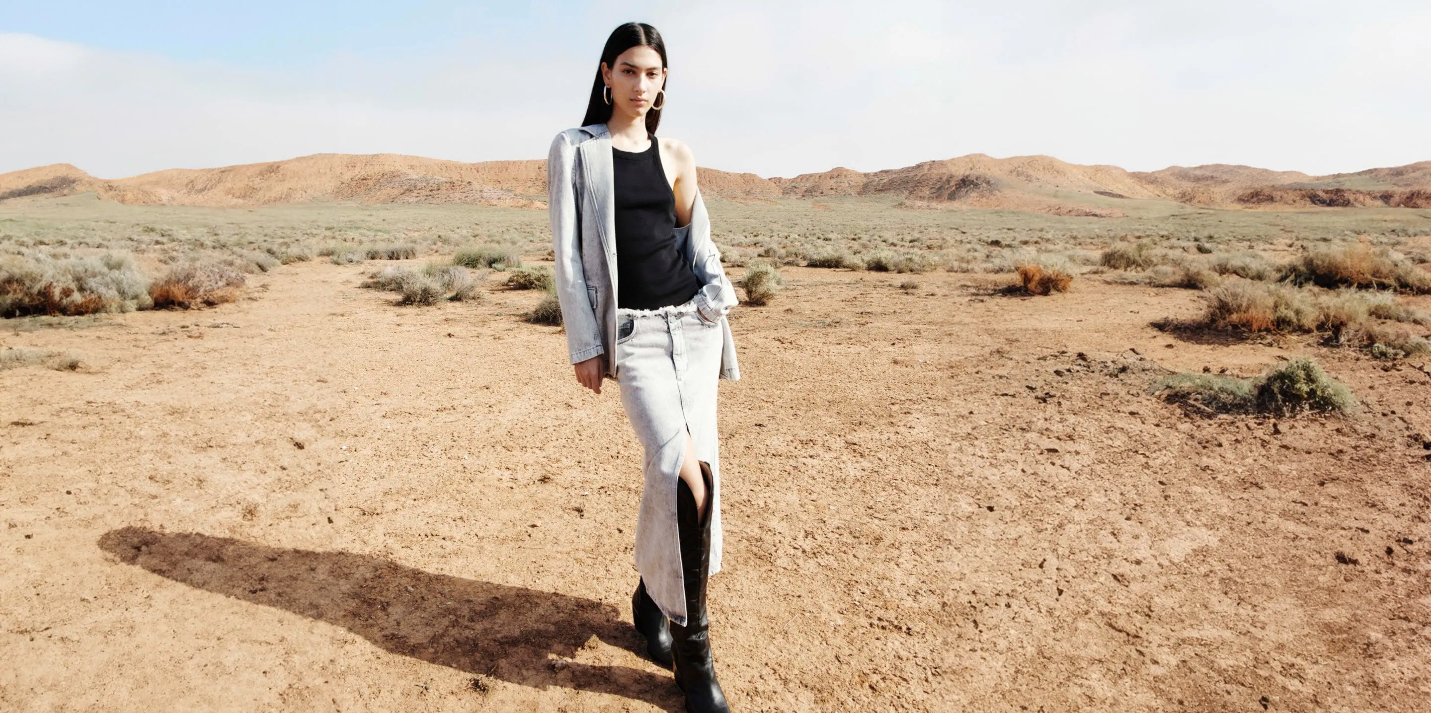 Photograph showing a female model wearing a light denim jacket, light denim skirt and black vest standing in the desert.