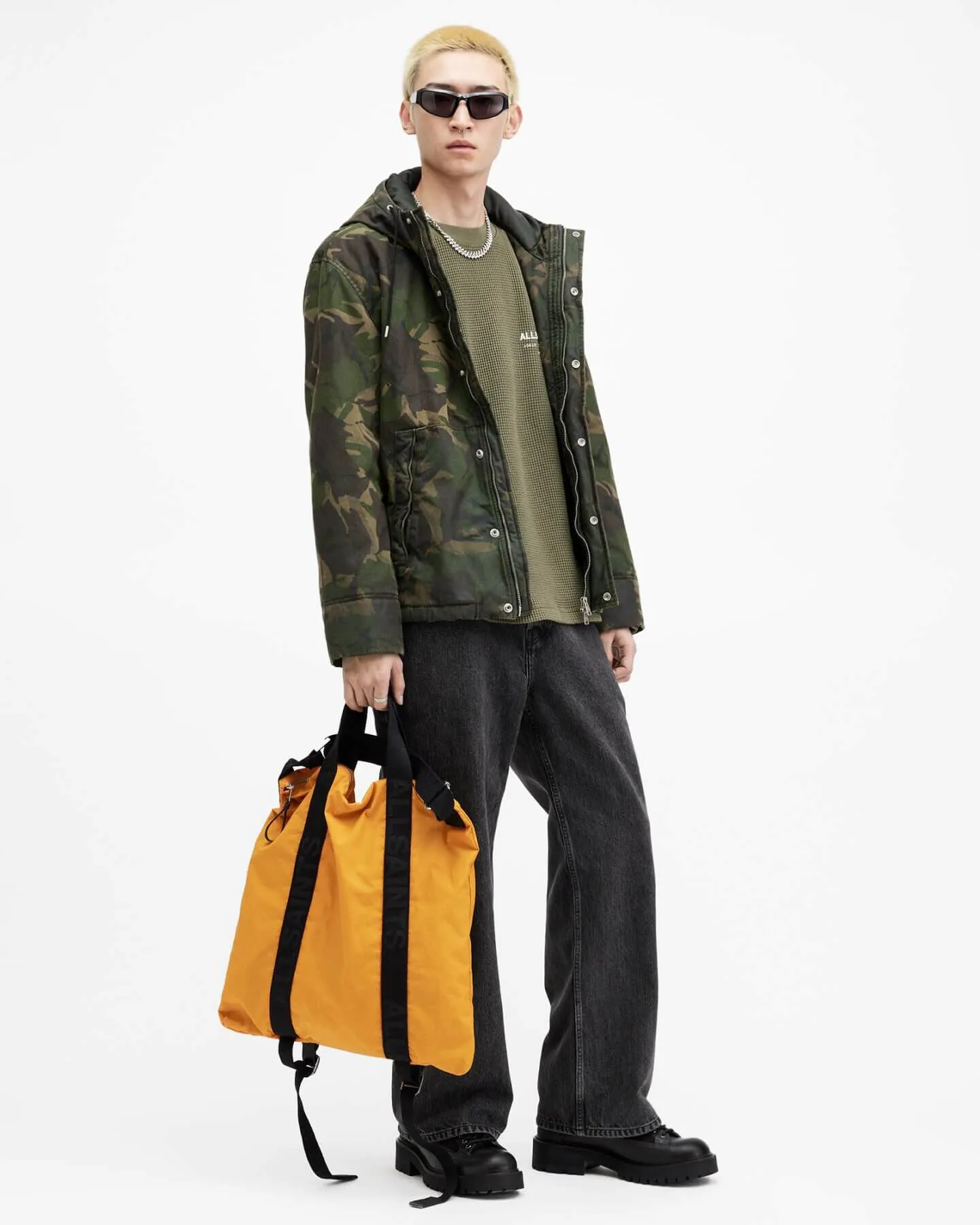 Man wearing a camouflage print jacket over a green t-shirt, with grey wide leg jeans and holding an orange backpack.