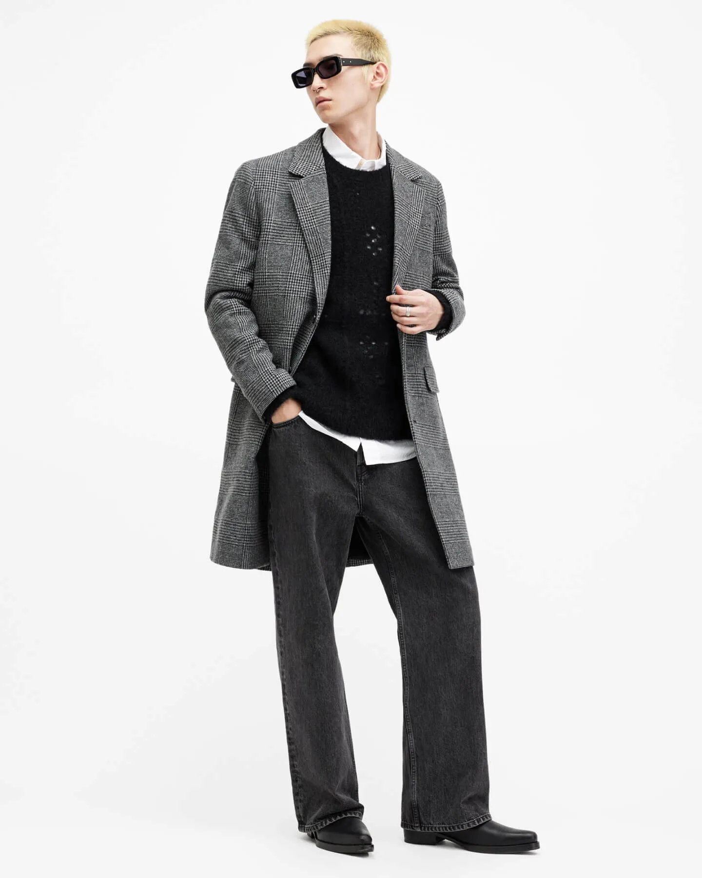 A man wearing a grey houndstooth coat over a black holey jumper, with grey wide leg trousers and black sunglasses.